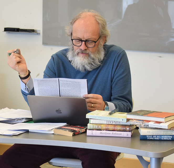 Paul Doerksen, man with full white beard and glasses, reading from pages and stacks of Mennonite books beside him