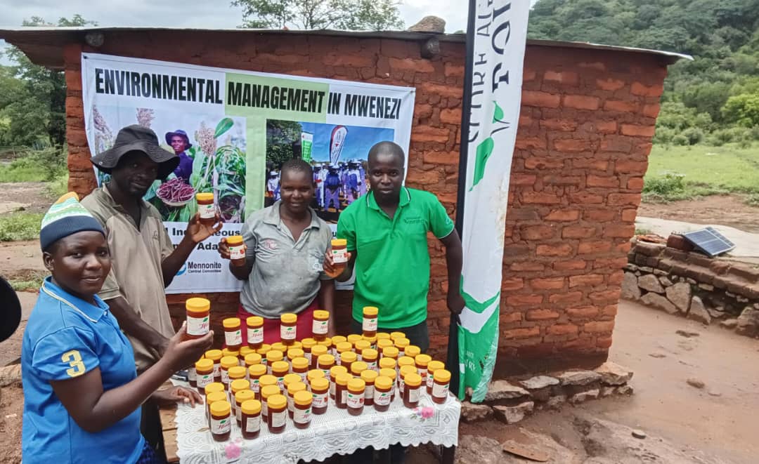 Family members proudly showcasing honey for sale next to Score Against Poverty staff member, surrounding bottles of honey laid out on a table outdoors.