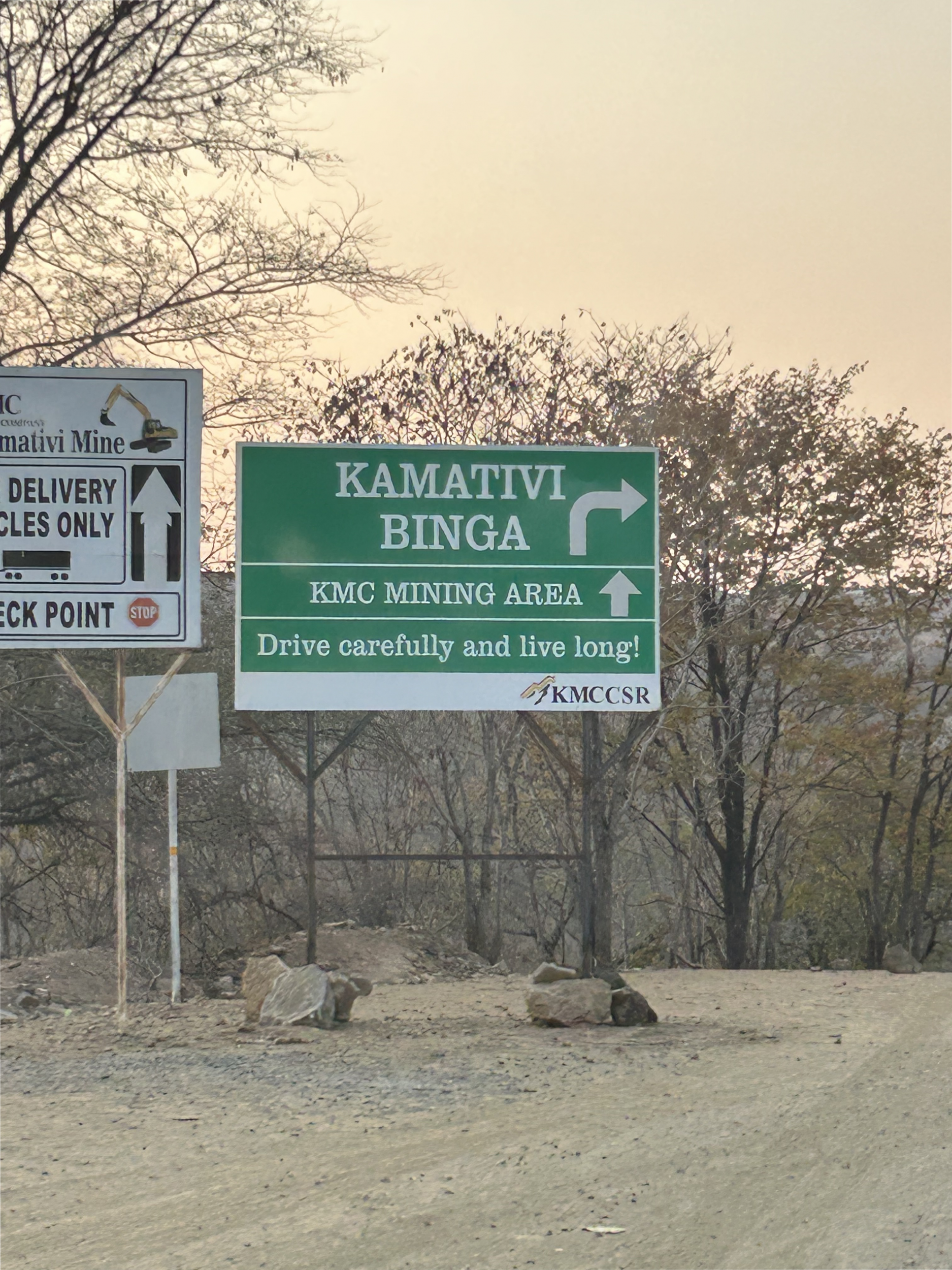 A green directional road sign that reads "Kamativi Binga, KMC mining area, drive carefully and live long!"